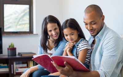 Happy multiethnic family sitting on sofa together looking at daughter's drawing book. Black father reading the homework on exercise book of his cute kid. Parents feeling proud on progress of daughter.